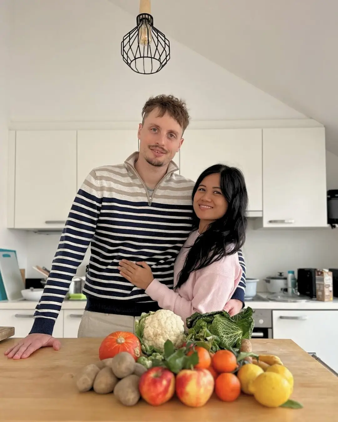 A couple stands in a bright, minimalist kitchen, smiling and hugging behind a wooden counter filled with colorful fresh produce like apples, potatoes, cauliflower, pumpkin, leafy greens, lemons, and mandarins.